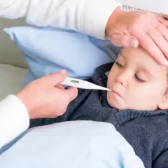 Dziecko leży w łóżku, a opiekunka mierzy mu temperaturę. Dłoń opiekunki delikatnie dotyka czoła malucha, podczas gdy druga trzyma termometr. To obraz troski i dbania o zdrowie, przypominający o tradycyjnych metodach, jak stawianie baniek u dzieci.