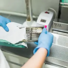 Ręce w niebieskich rękawiczkach wkładają tackę ze sterylnymi narzędziami do urządzenia, pokazując, jak działa autoklaw.