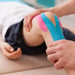 Fizjoterapeuta pokazuje, jak przykleić tejpy na kolano dziecka. Kolorowe plastry zapewniają wsparcie.