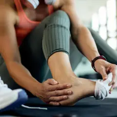 Kobieta w siłowni masuje kostkę, odczuwając piekący ból po treningu. Obok leżą buty sportowe.