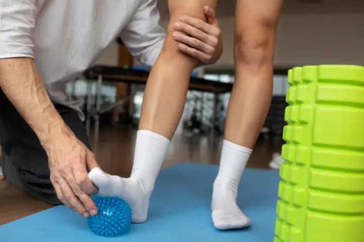 Fizjoterapeuta pomaga w rehabilitacji stawu skokowego, wykonując ćwiczenia z użyciem niebieskiej piłeczki do masażu.