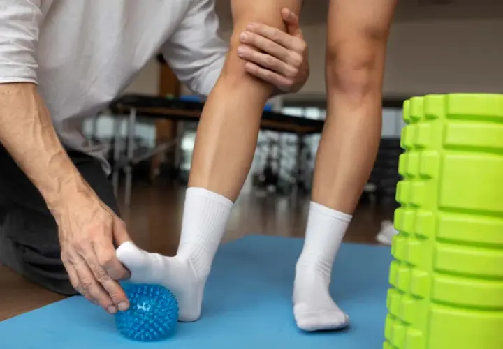 Fizjoterapeuta pomaga w rehabilitacji stawu skokowego, wykonując ćwiczenia z użyciem niebieskiej piłeczki do masażu.