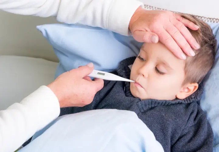 Dziecko leży w łóżku, a opiekunka mierzy mu temperaturę. Dłoń opiekunki delikatnie dotyka czoła malucha, podczas gdy druga trzyma termometr. To obraz troski i dbania o zdrowie, przypominający o tradycyjnych metodach, jak stawianie baniek u dzieci.