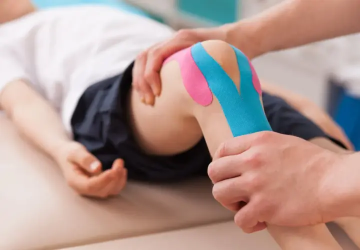 Fizjoterapeuta pokazuje, jak przykleić tejpy na kolano dziecka. Kolorowe plastry zapewniają wsparcie.