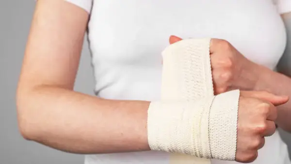 Osoba owija rękę bandażem. Widoczne są objawy stłuczenia nadgarstka: ból, obrzęk i ograniczenie ruchomości.